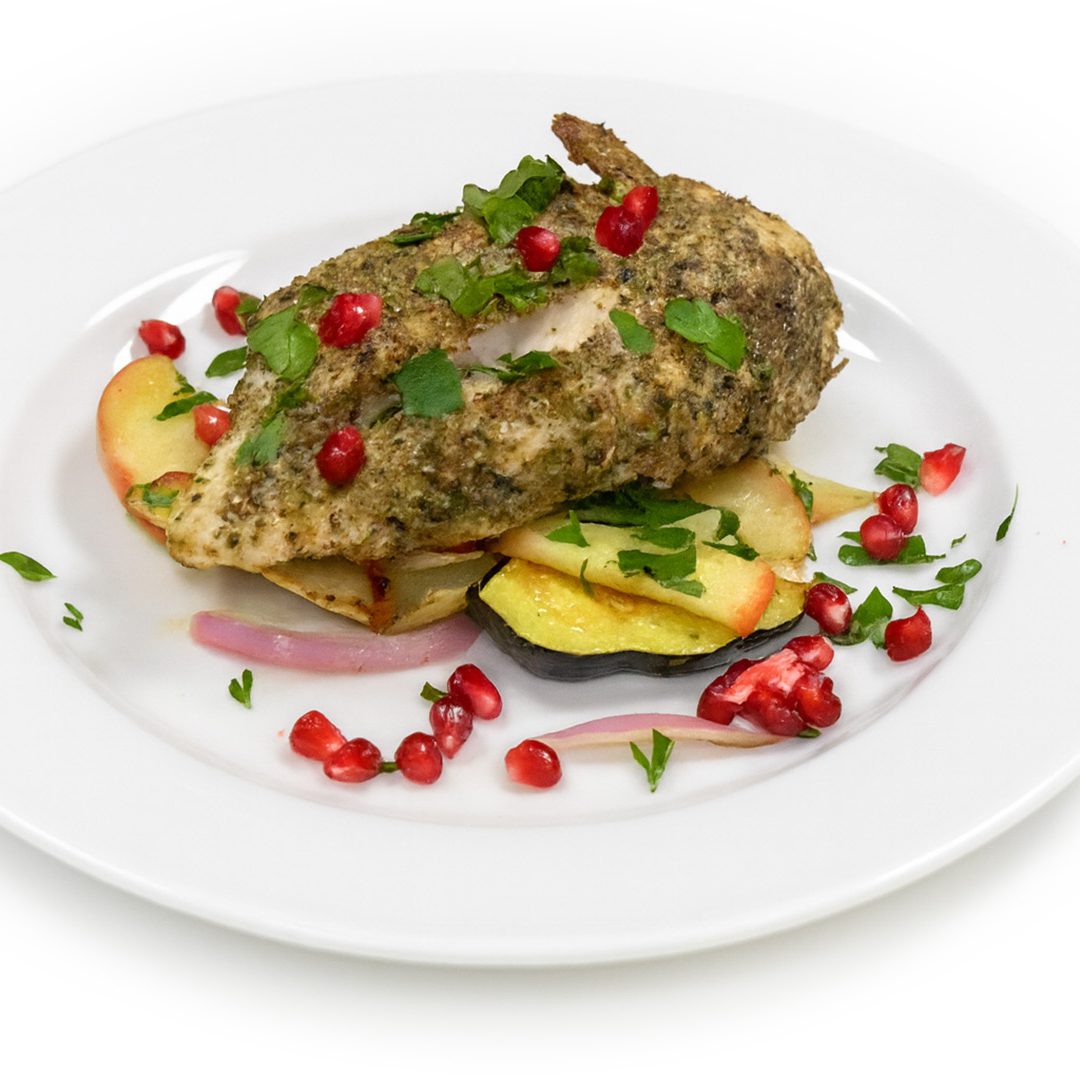 Overhead view of Herb-crusted chicken breast served on roasted apple, squash, and red onion slices, garnished with fresh herbs and pomegranate seeds on a white plate